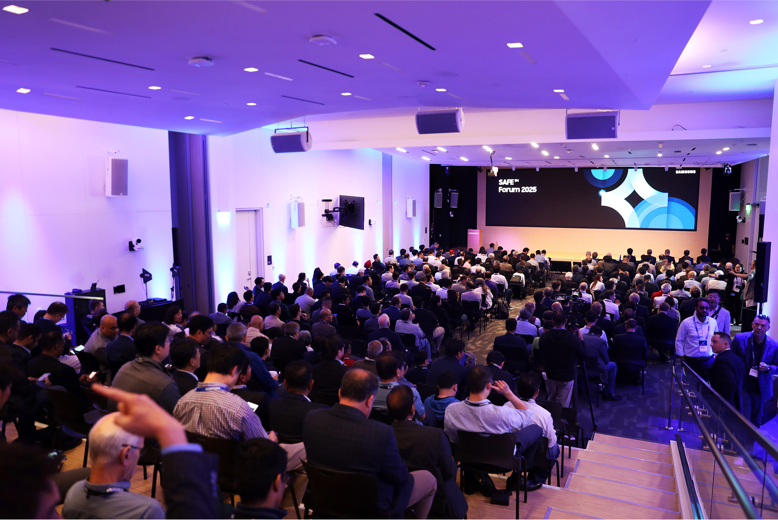 A large audience attentively listens during the Samsung SAFE Forum 2025, held in a modern, well-lit conference hall with a wide digital screen at the front.