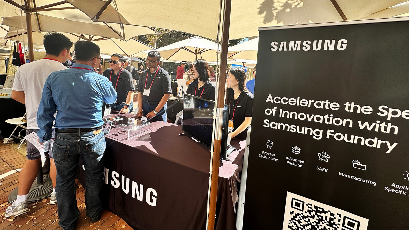 Samsung Foundry booth at a tech event with staff engaging visitors, with a large promoting banner.