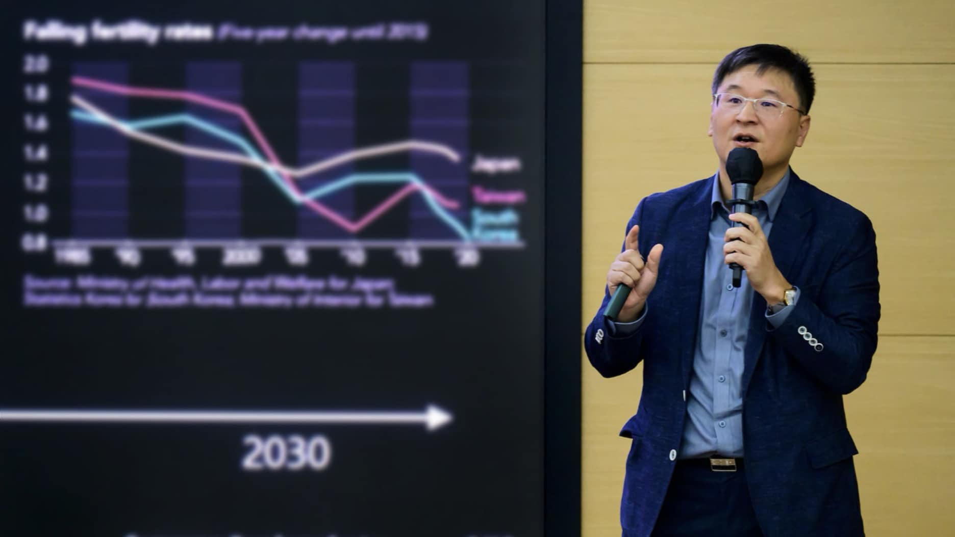 A presenter at the 2024 Semiconductor Tech Seminar speaking while referencing a slide showing trends in fertility rates over time.