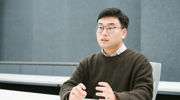 회의실에서 책상 앞에 앉아 손을 약간 올린 채 이야기하는 남성.