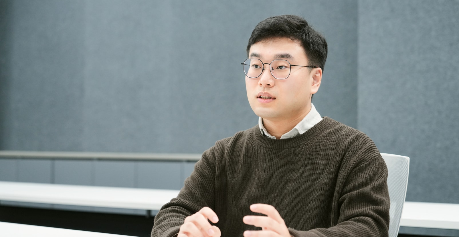 회의실에서 책상 앞에 앉아 손을 약간 올린 채 이야기하는 남성.