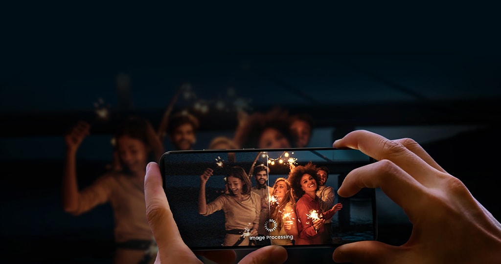 A person holds a smartphone capturing a group of people celebrating at night, emphasizing the phone's low-light photography and image processing capabilities.