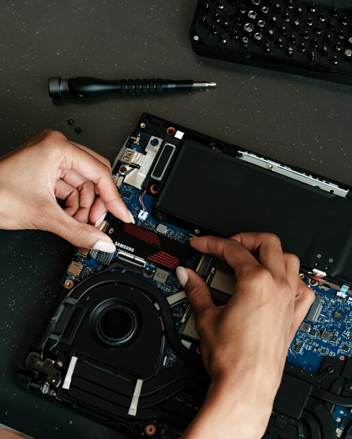 A person carefully placing a Samsung SSD into a laptop's internal slot, with a screwdriver and screws visible nearby, indicating a hardware upgrade process.