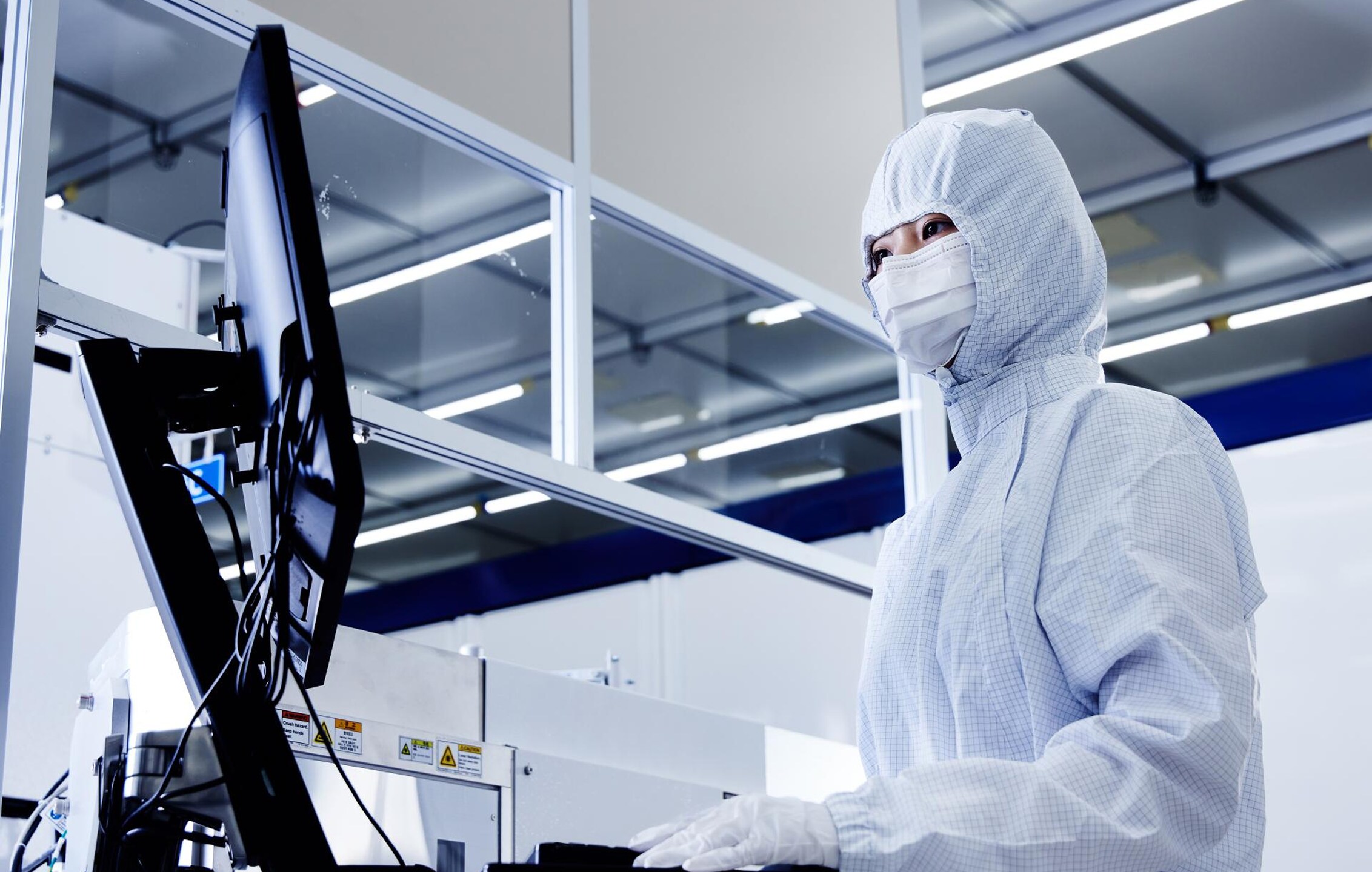 半導体工場のクリーンルーム内で、防塵服とマスクを着用した作業員がコンピュータ画面で製造工程データを監視している様子.