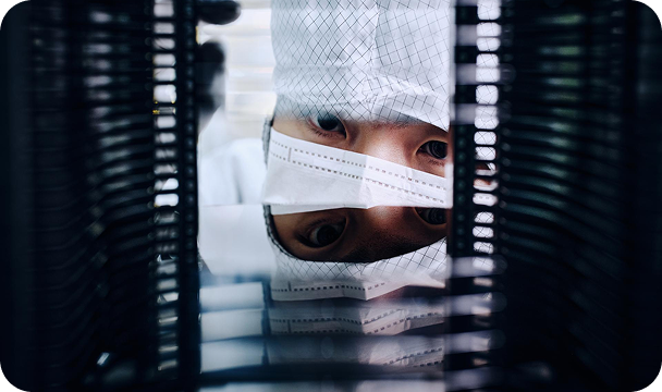 クリーンルームで防塵服を着用したエンジニアが装置ラック越しに半導体工程の状態を確認する様子