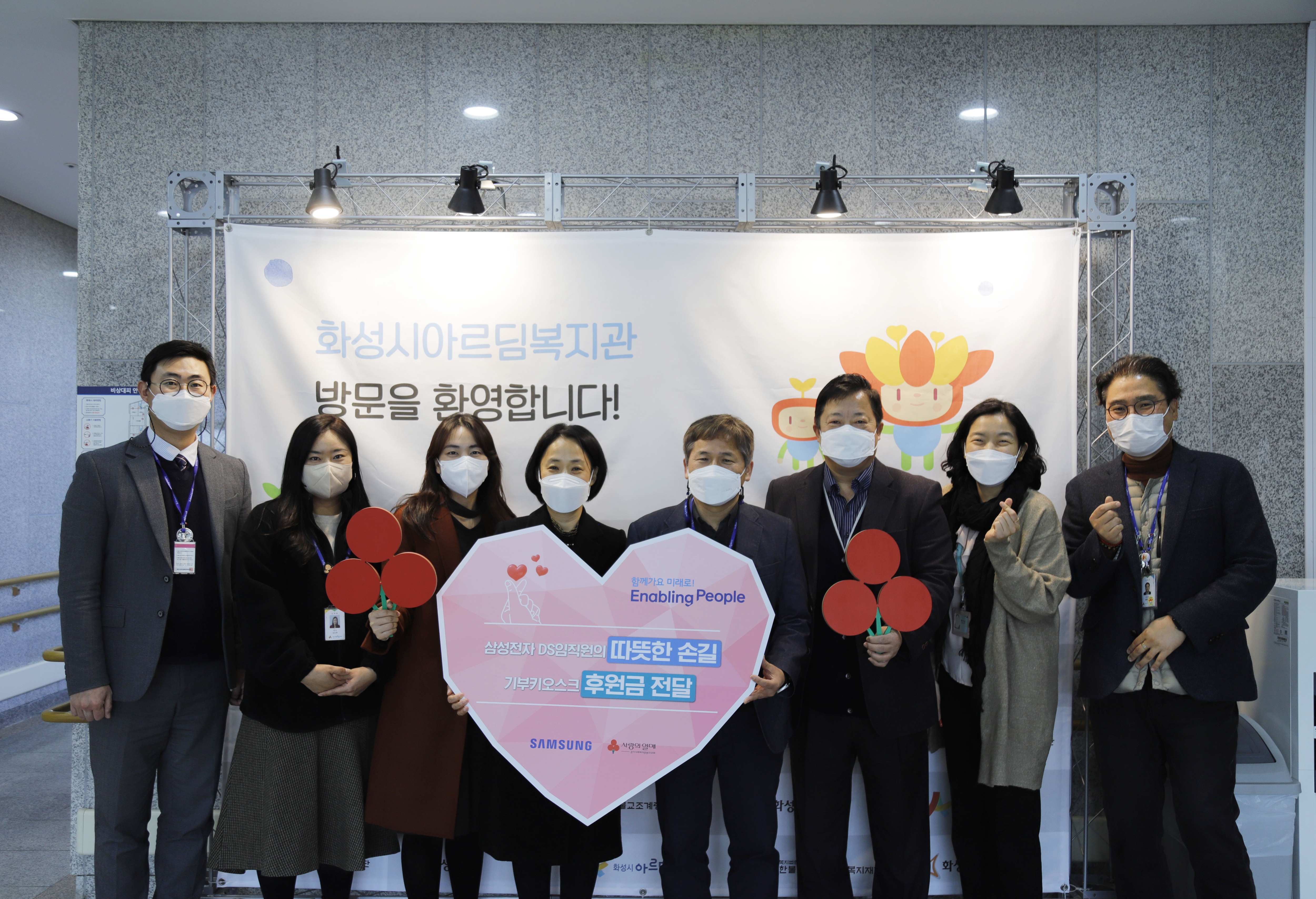 Samsung Semiconductor and Community Chest of Korea employees are holding up a sign together that's in the shape of a heart. The sign says that Samsung Semiconductor has donated money collected via donation kiosks to Community Chest of Korea