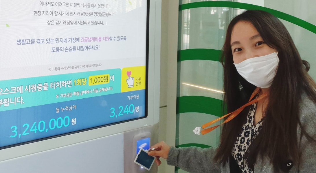 기부 키오스크에 마스크를 쓴 여직원이 사원증을 찍으며 앞을 보고 있는 모습입니다.