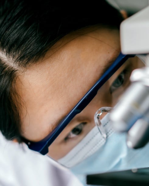 Close-up of person seen from the front, wearing safety goggles and a mask while looking at something on the table.