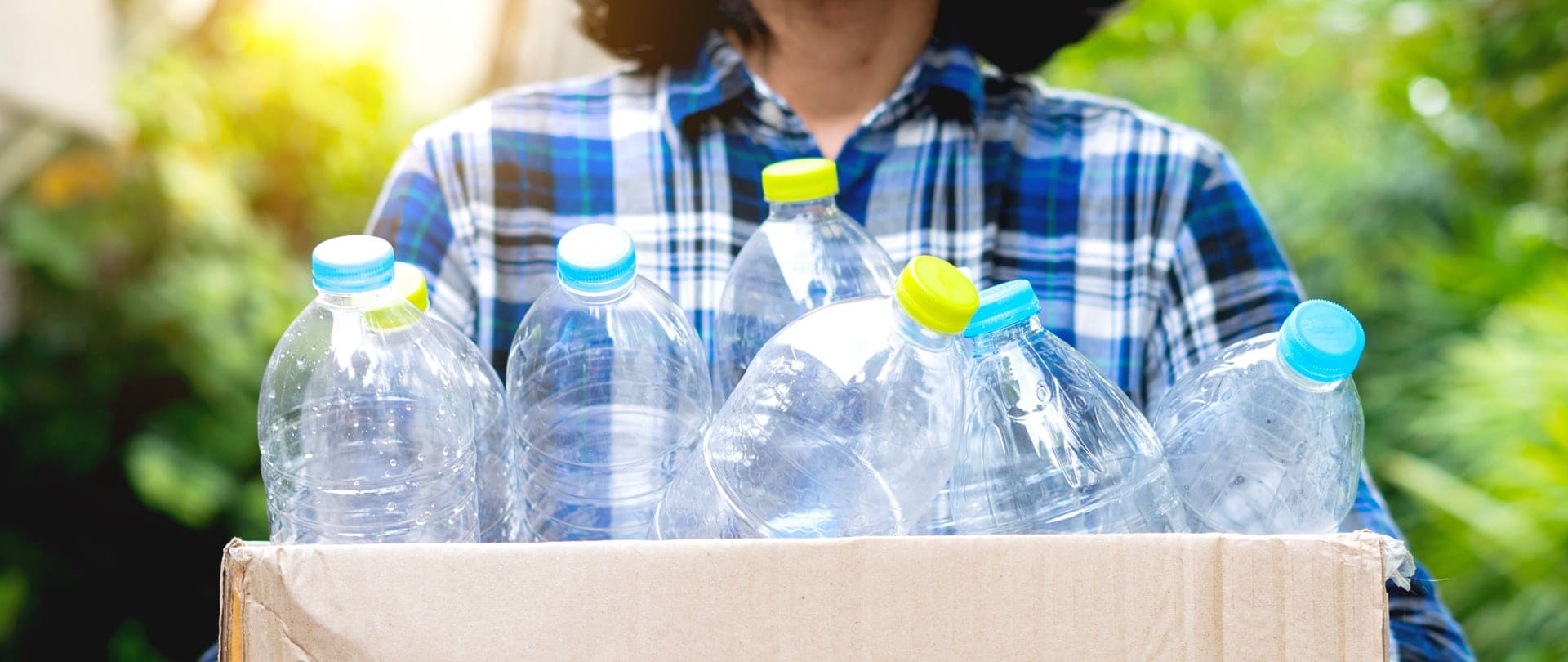 A woman wearing a blue checkered shirt is holding a box of several empty plastic bottles.