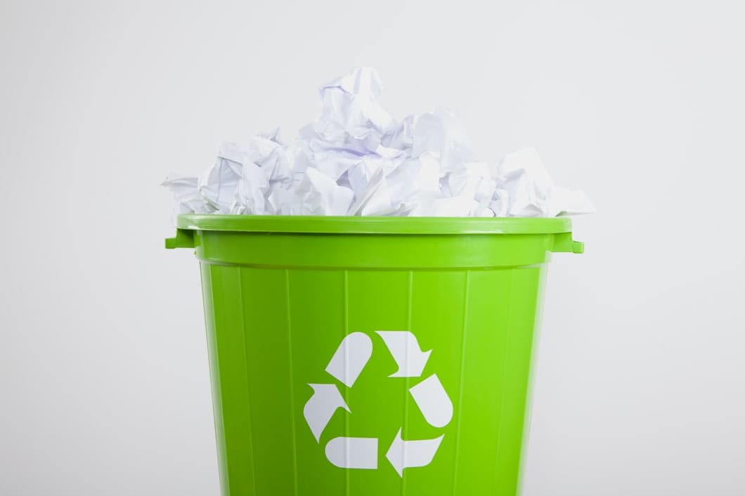 Crumpled white paper waste in a green trash can with a white recycling mark.