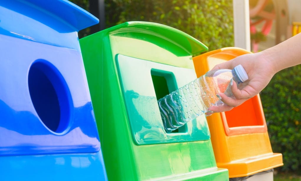 Blue, green, and orange recycling bins, with plastic being thrown in the green recycling bin.