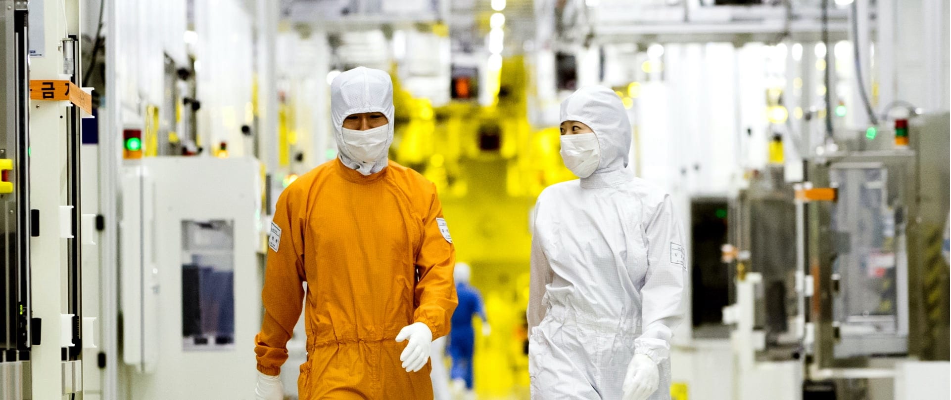 Two people walking in Samsung's semiconductor manufacturing plant, one in orange protective gear and one in white.